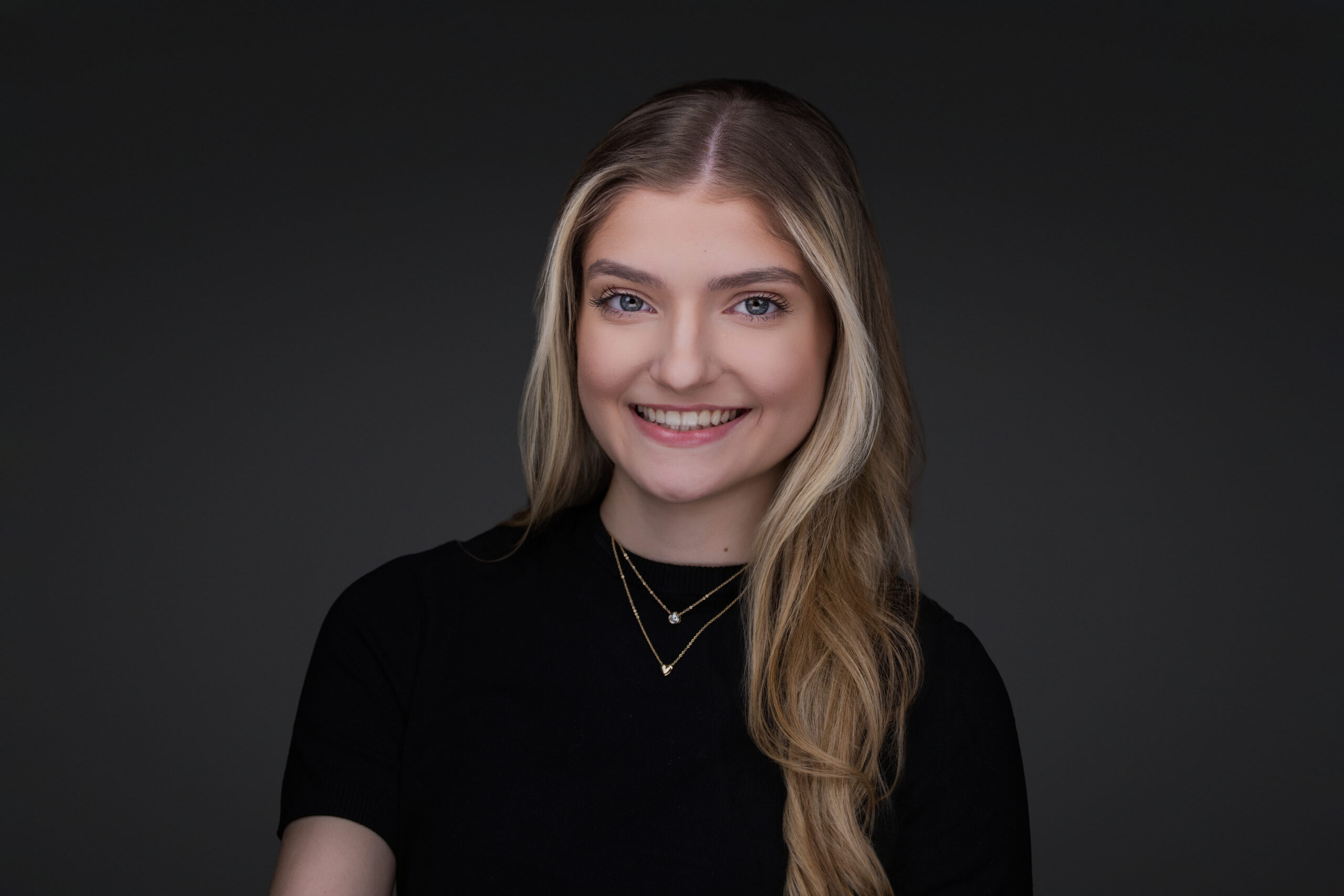 A portrait of class of 2026 Senior Isabella in a black top on a grey background.